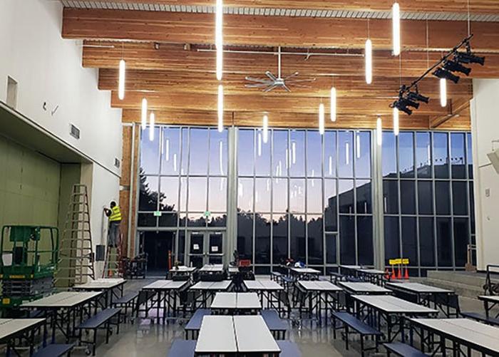 a large room with a wall of windows and exposed wood beams a stage is on the left and there are lunch tables