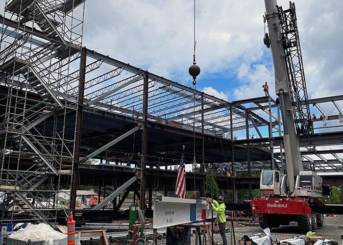 a crane line is being attached to a white steel beam with red and blue stripes. there is a US flag and an evergreen tree attached to the top of the beam